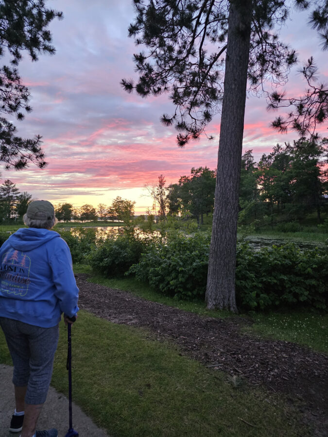 A woman with a cane walking near a pond with a sunset in the sky.